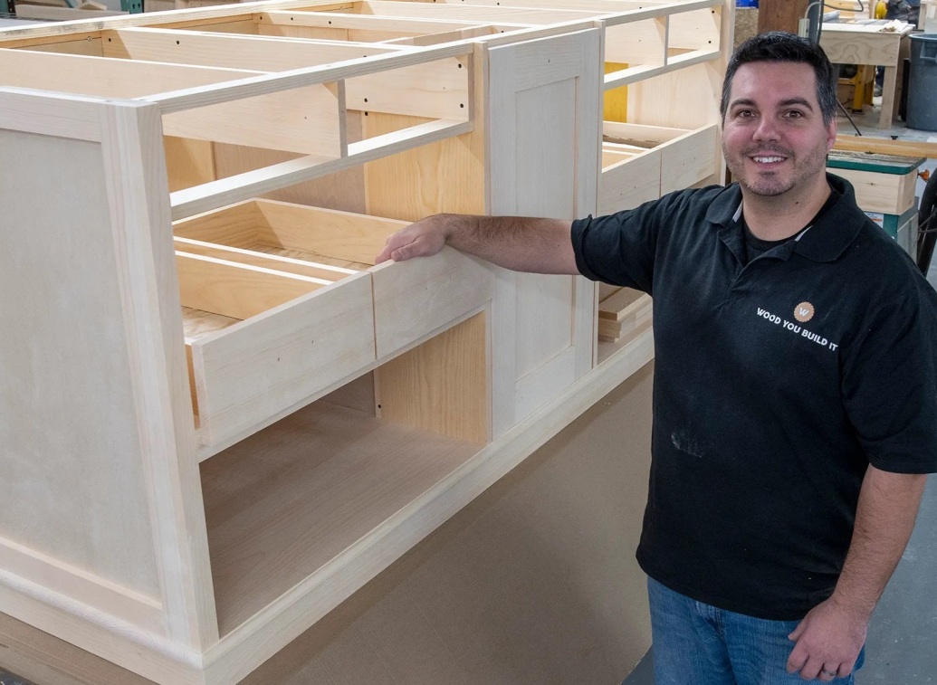 Justin Robichaud, founder of Wood You Build It, standing next to a large unfinished custom kitchen island in the workshop.